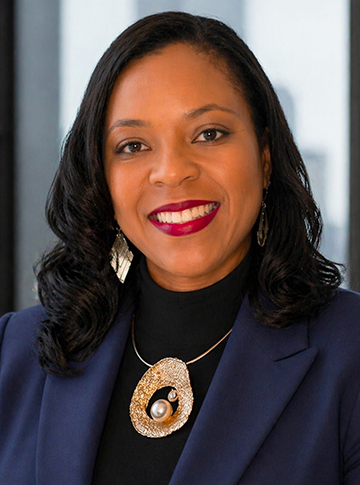 A smiling woman with long black hair wearing a dark blue blazer and a large gold necklace.