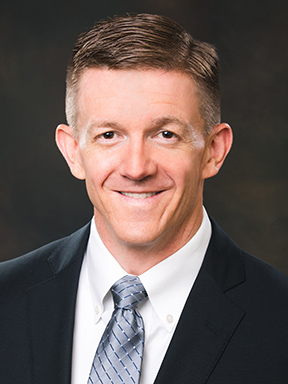 A smiling man in a dark suit wearing a white dress shirt and a gray patterned necktie.