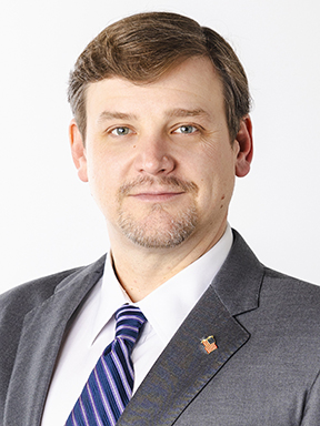 A man with facial hair in a gray suit wearing a white dress shirt and blue striped necktie.