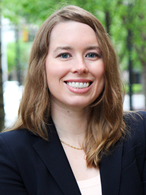 A young woman with long hair wearing a dark blazer with a light top smiles with trees in the background.