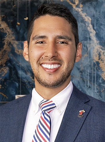 Young hispanic man smiling and wearing a blue suit, white dress shirt, and red, white and blue striped necktie.