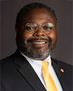 A man in a dark suit and a white dress shirt wearing a yellow necktie.